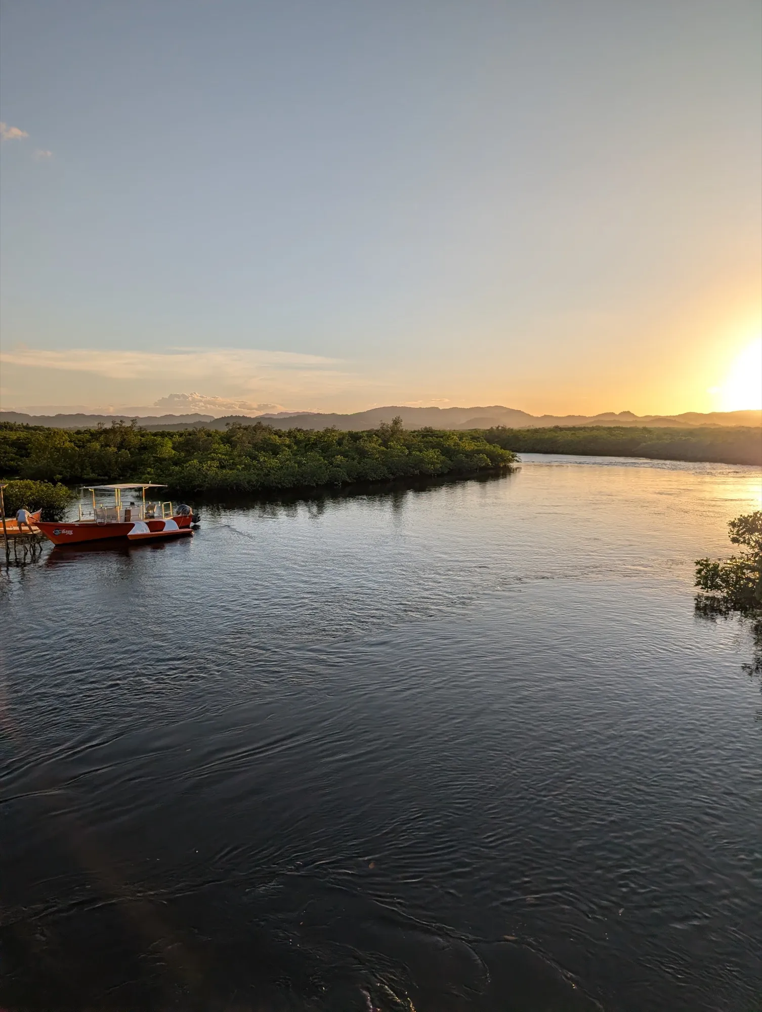 Sunset over a mangrove estuary in Siargao with a small wooden boat moored at a dock — typical scene from the Del Carmen area along the western route