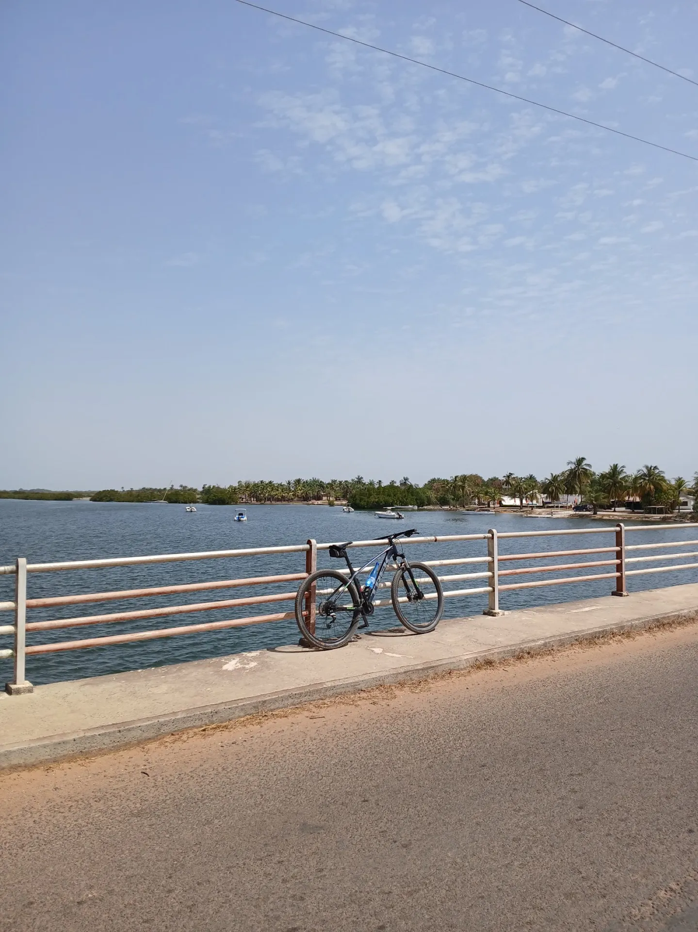Mountain bike resting against the white-and-red bridge railing over a calm Siargao lagoon with palm-fringed shore and small fishing boats